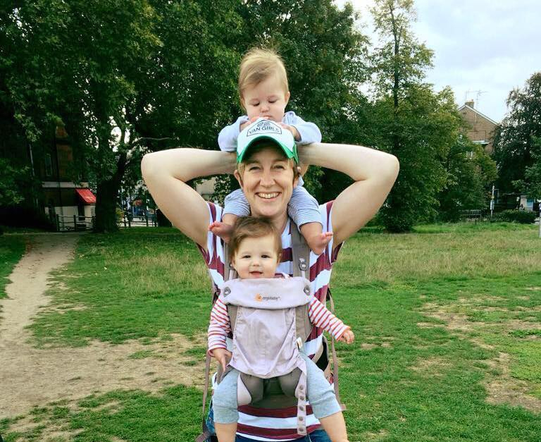 Woman in a park with one baby strapped to her front and another baby on her shoulders.